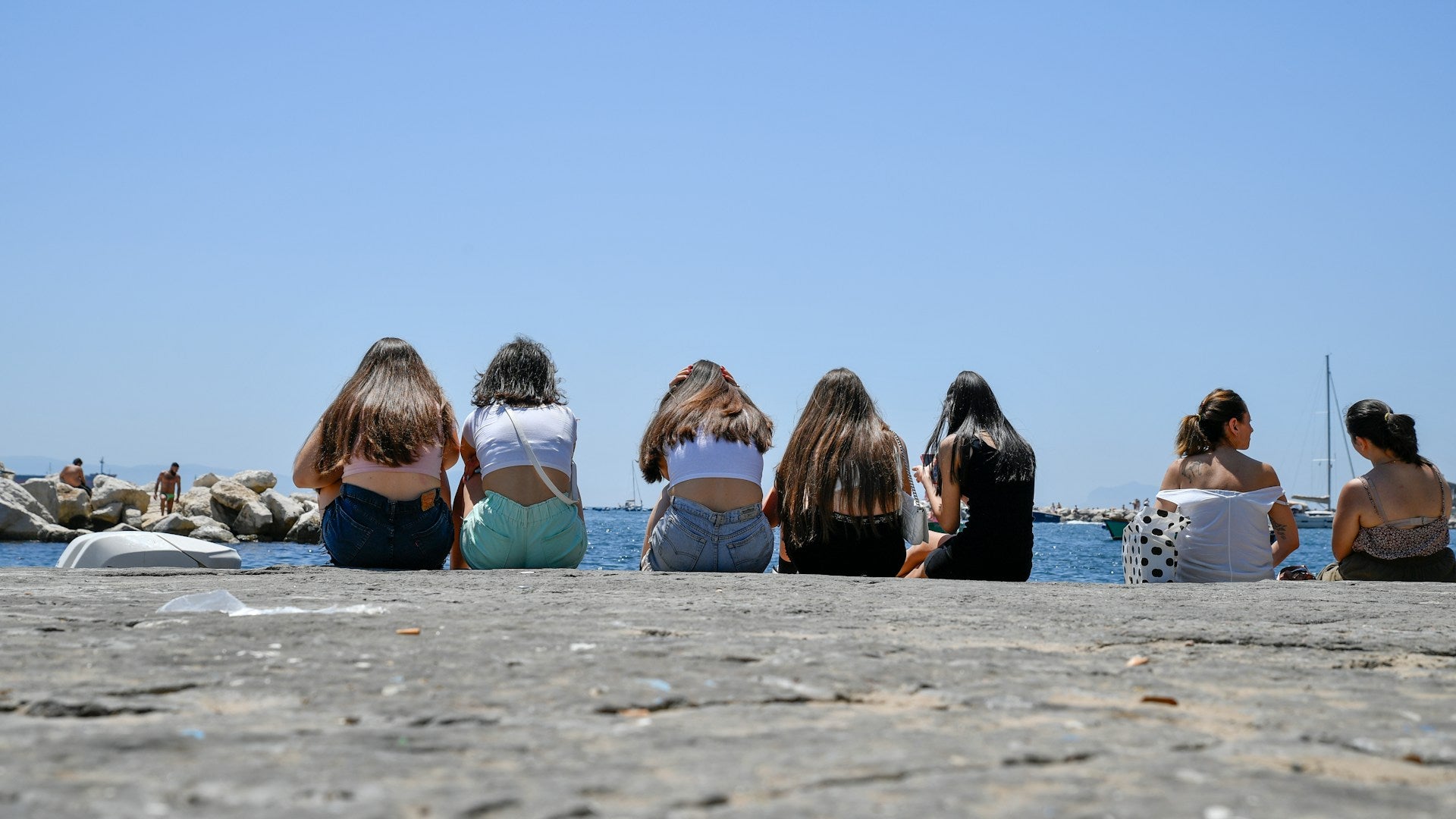 A group of women sitting together on a waterfront pier, backs to the camera, looking out at the sea